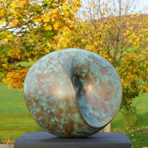 Abstract, round bronze sculpture, green-brown patinated, in front of an autumn landscape. goldleuchtender Siliziumbronze.